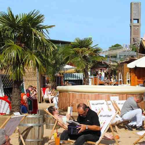 Gäste entspannen in einem Beach Club mit Sand, Palmen und Liegestühlen.