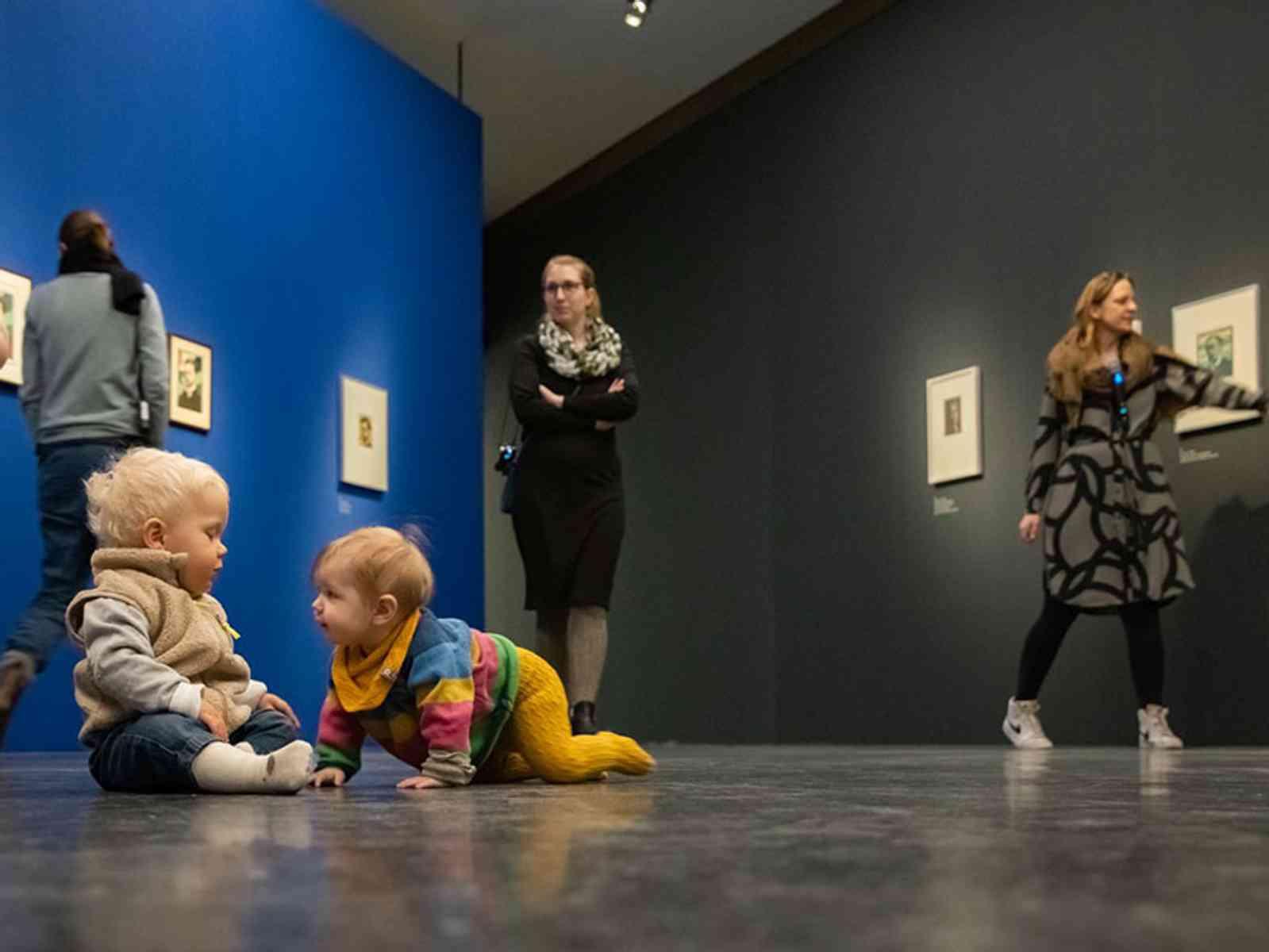 Kinder spielen auf dem Boden, während Eltern Kunst anschauen