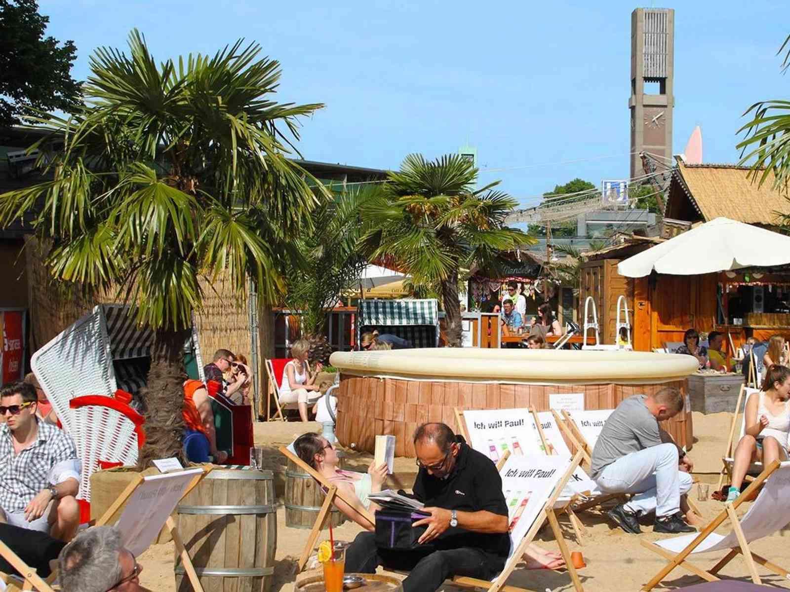 Gäste entspannen in einem Beach Club mit Sand, Palmen und Liegestühlen.