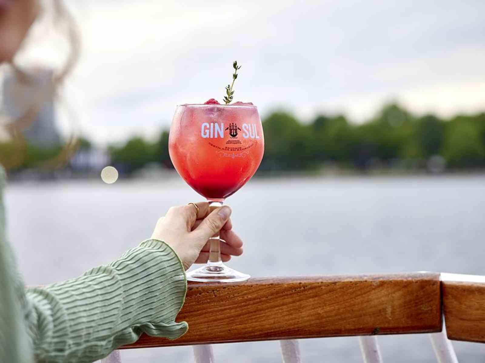 Person hält ein Gin Glas in die Kamera. Im Hintergrund liegt die Alster.