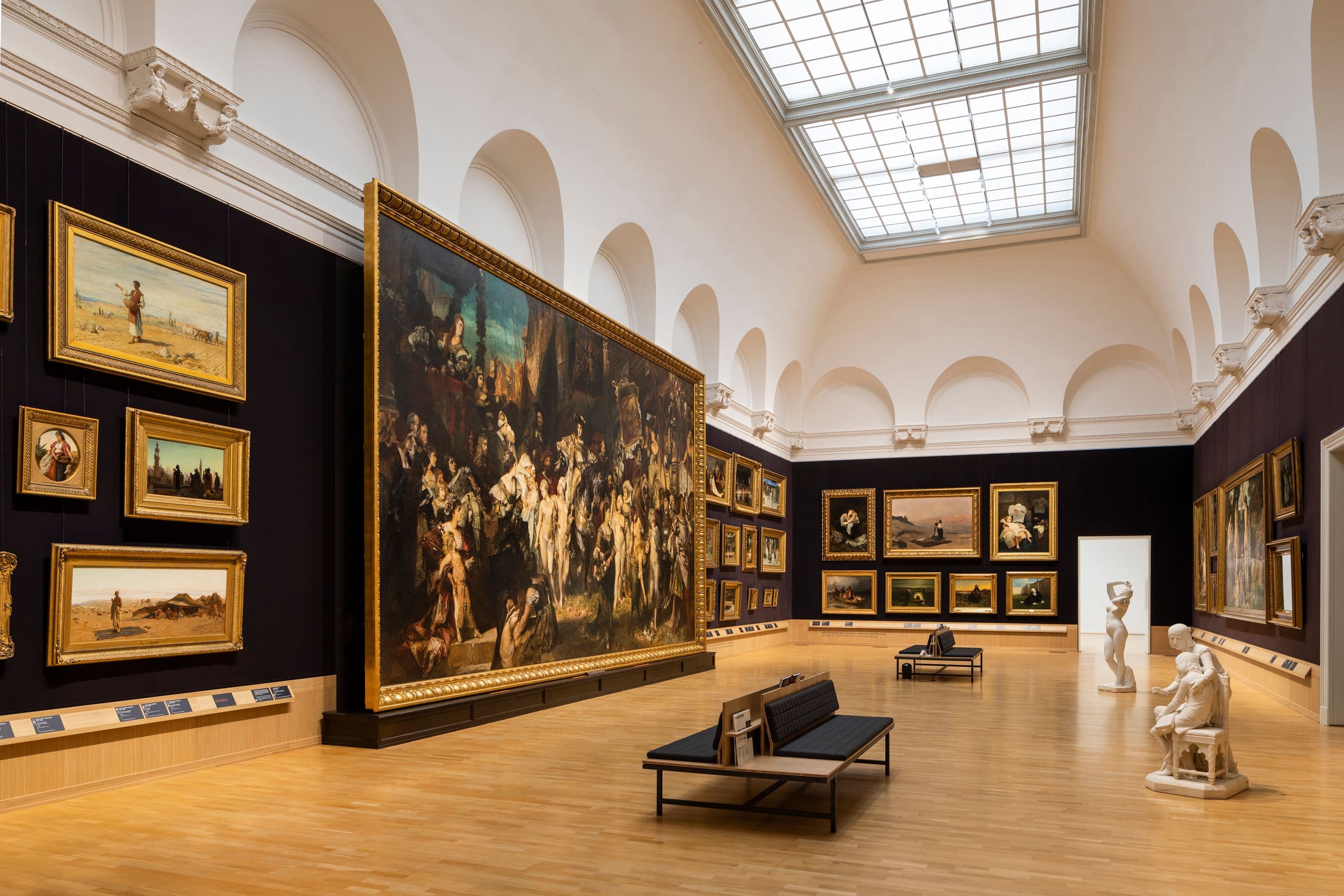Raumansicht des Makart-Saal der Hamburger Kunsthalle. An allen Wänden hängen kleinere Gemälde und ein ganz großes. Dabei handelt es sich um Hans Makarts "Einzug Karls V.s in Antwerpen" aus dem Jahr 1878. In der Raummittel stehen jeweils zwei Skulpturen und Sitzbänke.