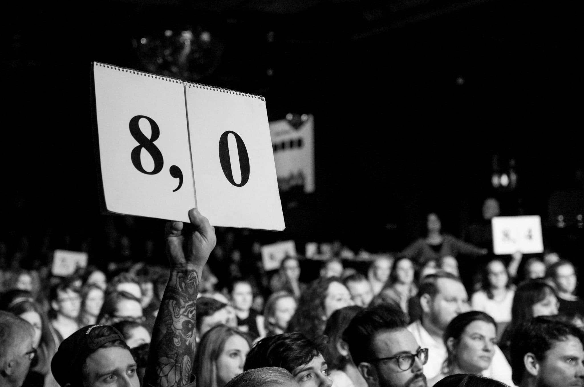 Schild 8,0 wird von einer Person in einem Publikum hochgehalten.