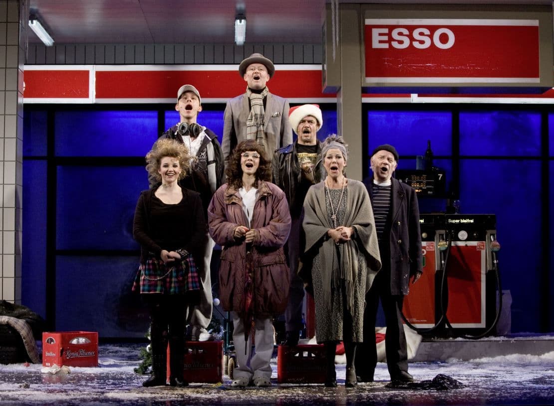 Eine Gruppe Schauspieler:innen stehen vor einer Tankstelle und singen gemeinsam