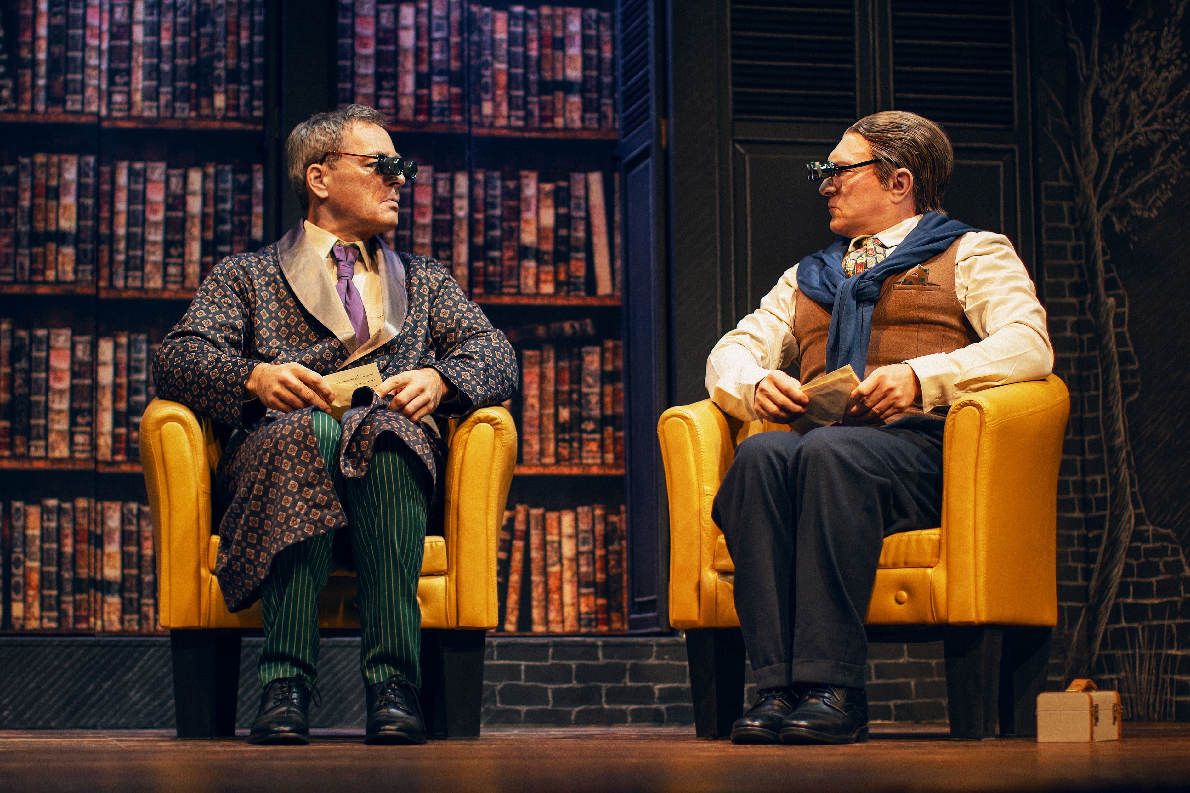 Bühnenbild des Sherlock Holmes Theaterstücks. Zwei Männer sitzen auf Sesseln und tragen lupenartige Brillen.