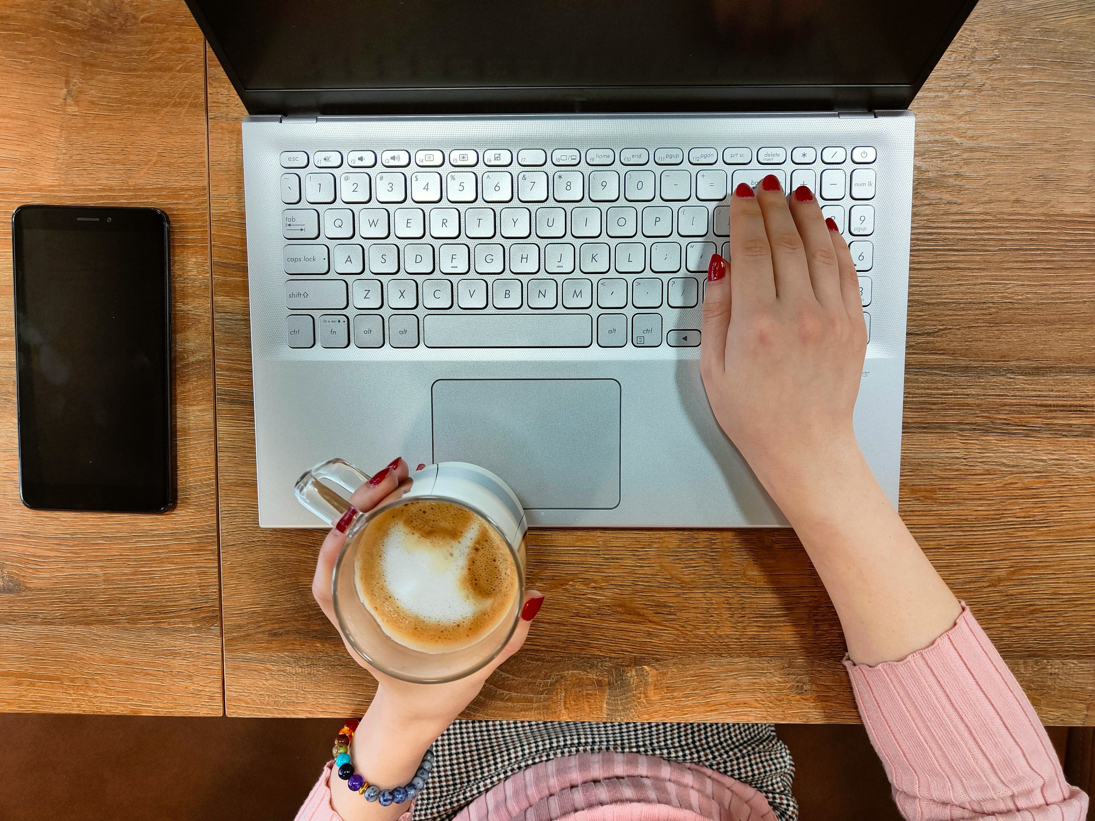 Eine Person hält einen Kaffee in der Hand und tippt auf einem Laptop
