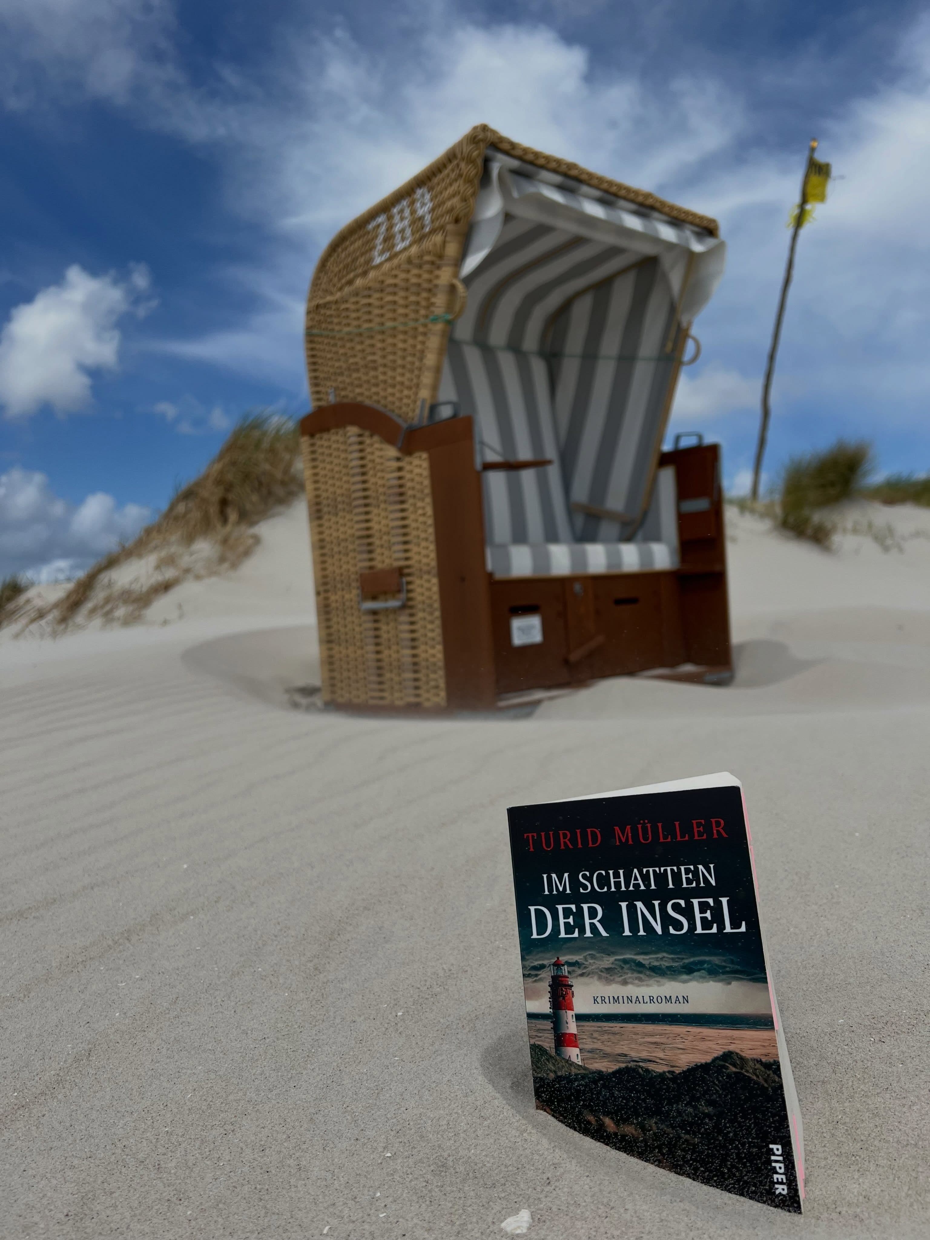 Turid Müllers Buch "Im Schatten der Insel" steckt im Sand und im Hintergrund steht ein Strandkorb