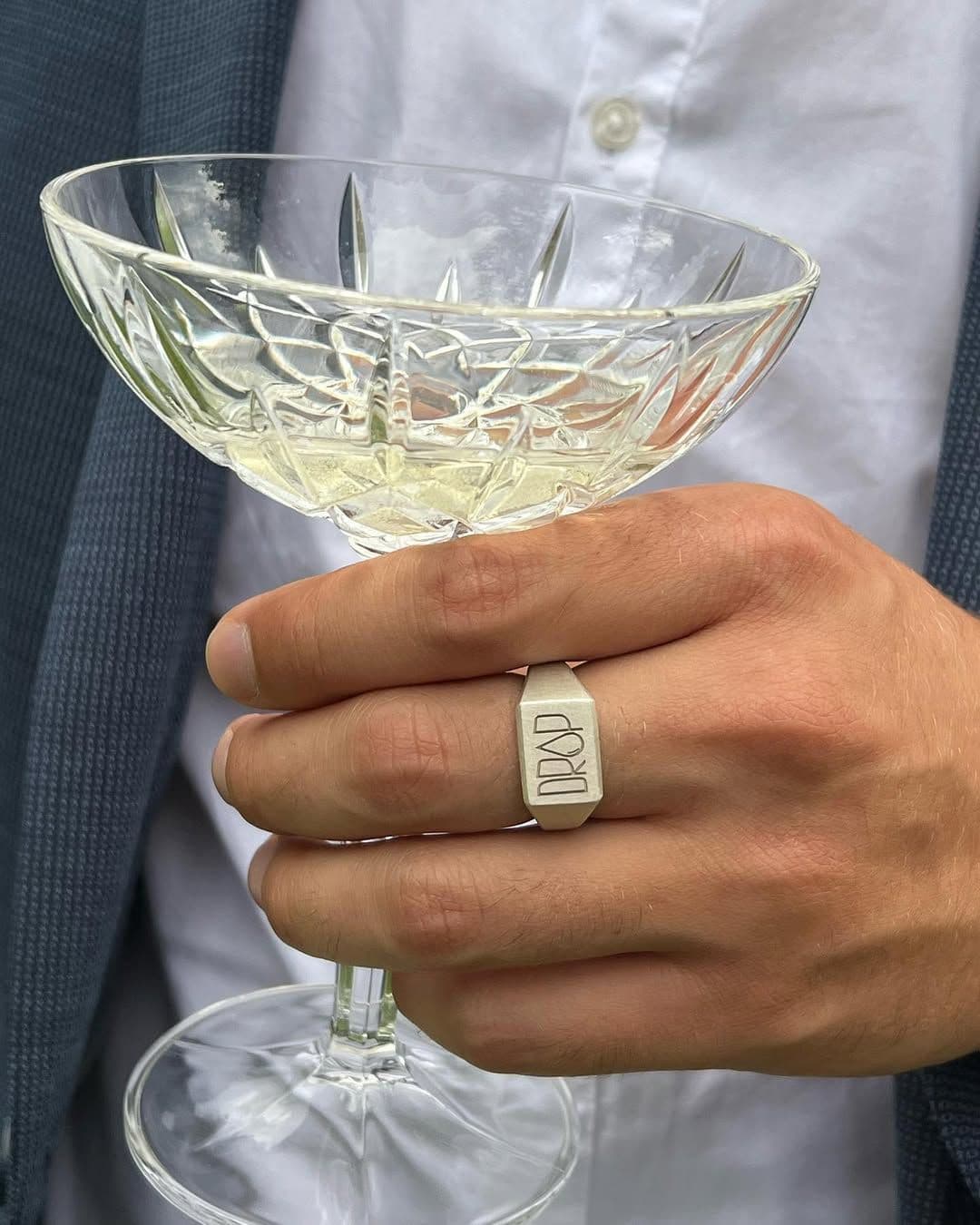 Nahaufnahme einer Hand mit Ring am Mittelfinger und einem Glas in der Hand