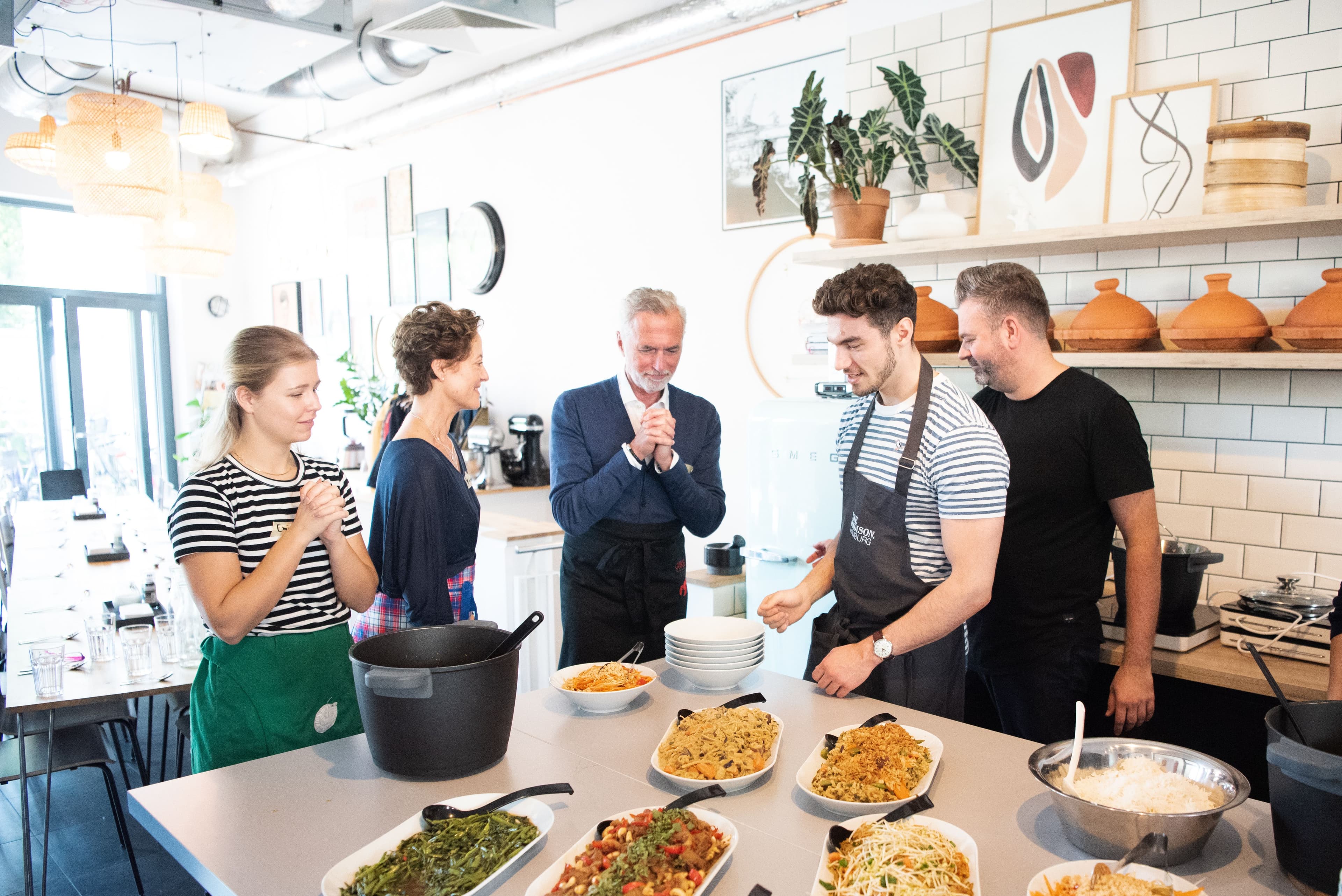 Fünf Menschen stehen an einer Kochinsel, auf der mehrere Teller und Schalen mit Essen stehen.