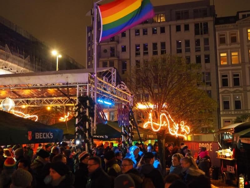 Blick auf den vollen Weihnachtsmarkt mit Getränkeständen, Bühne und einer gehissten Regenbogenfahne