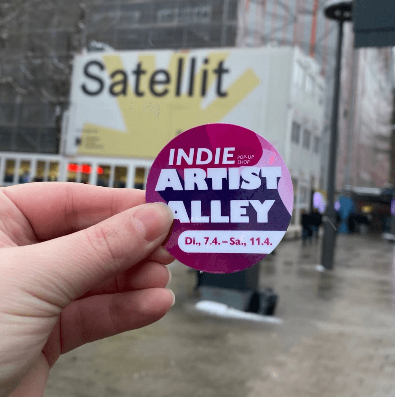 Hand hält Indie Artist Alley Sticker vor das Satellit Gebäude in Hamburg