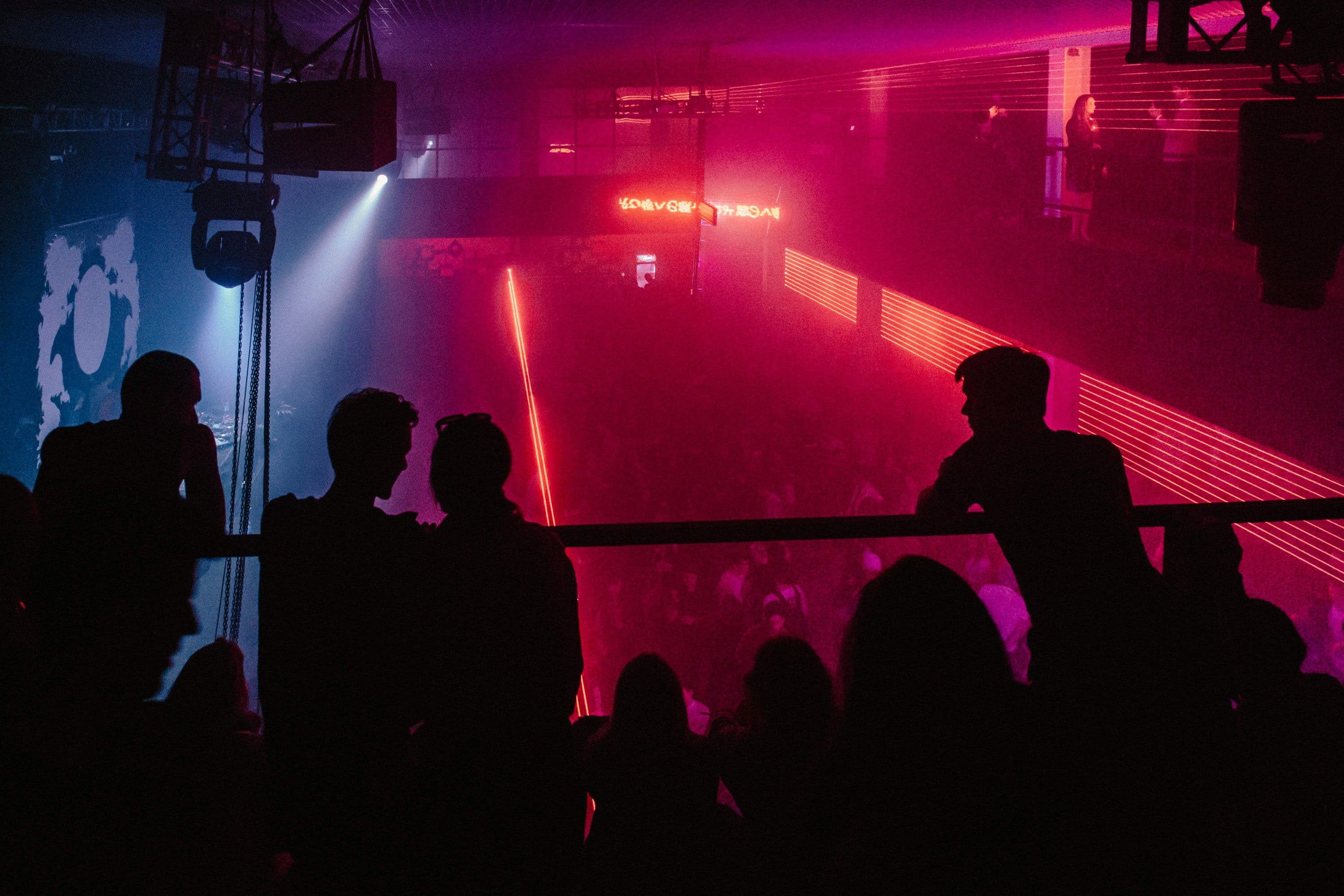 Menschen stehen in einem Club uns schauen auf die untere Etage herunter, wo ganz viele andere Menschen tanzen.