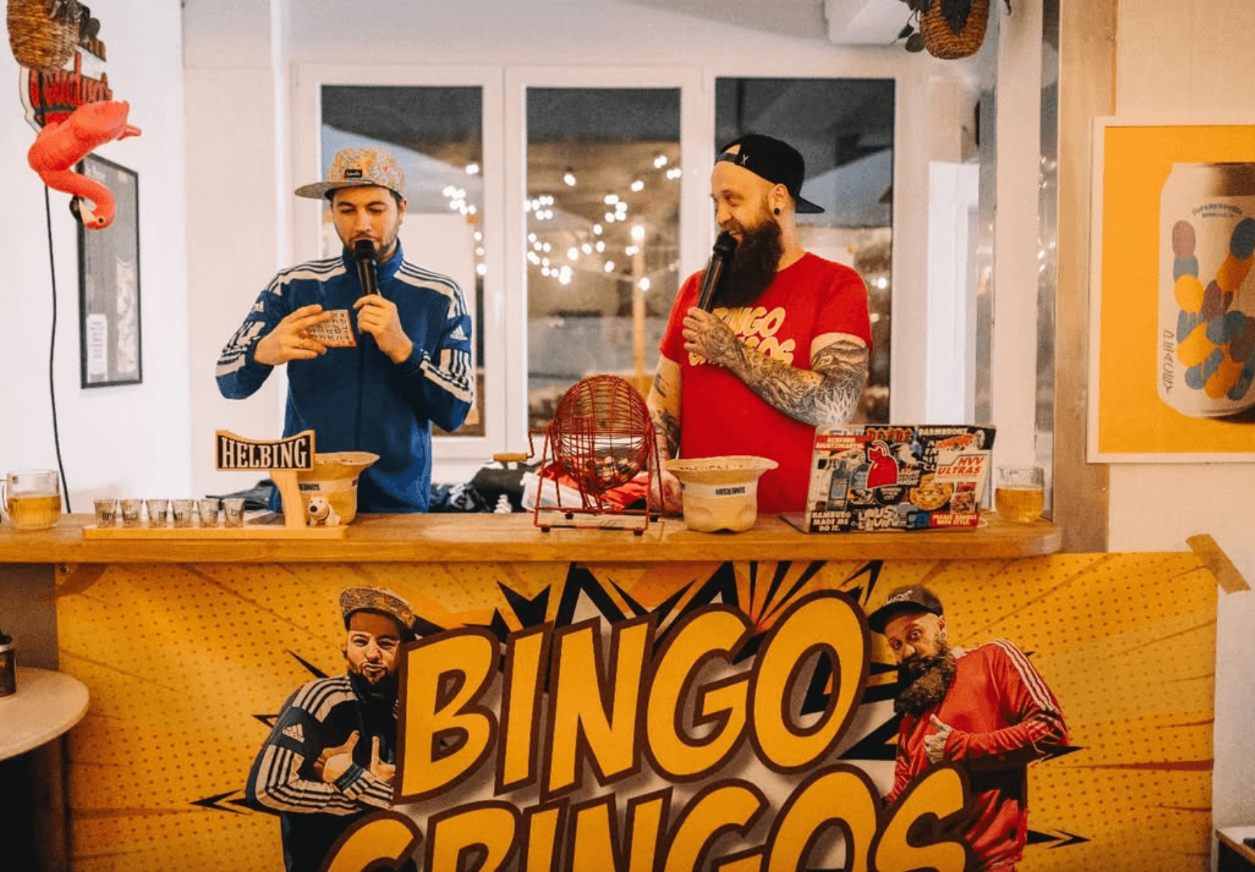 Die Beiden Mitglieder des Bingo-Duos Bingo Gringos stehen mit ihrem Bingo-Equipment und Mikrofonen in der Hand hinter einem Tresen.