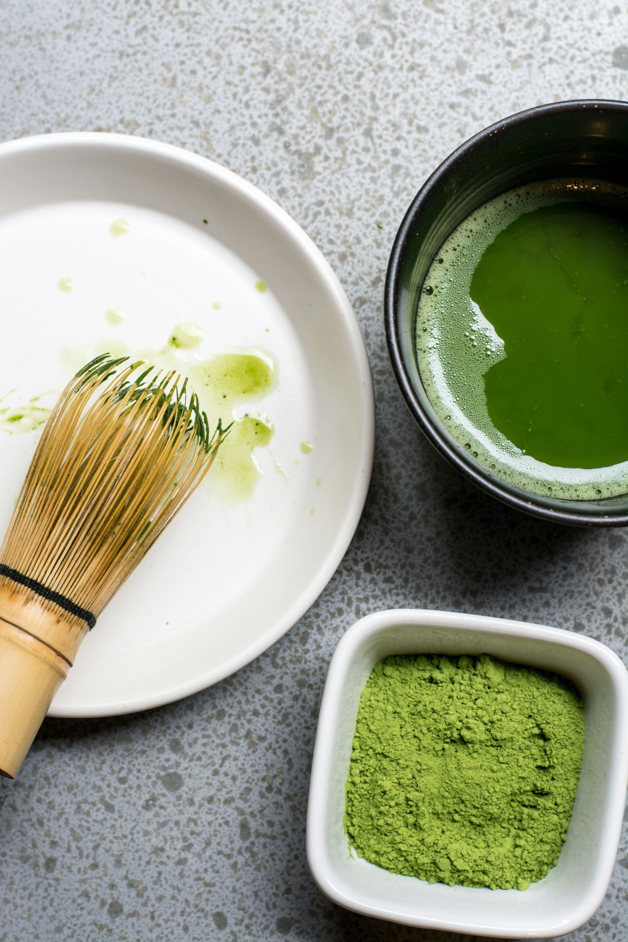 Grünes Matcha-Pulver in einem Gefäß, ein benutzter Matcha-Besen auf einem Teller und zubereiteter Matcha in einer Tasse