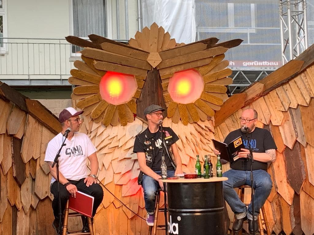 Drei Personen sitzen an Mikrofonen vor einer Eule aus Holz