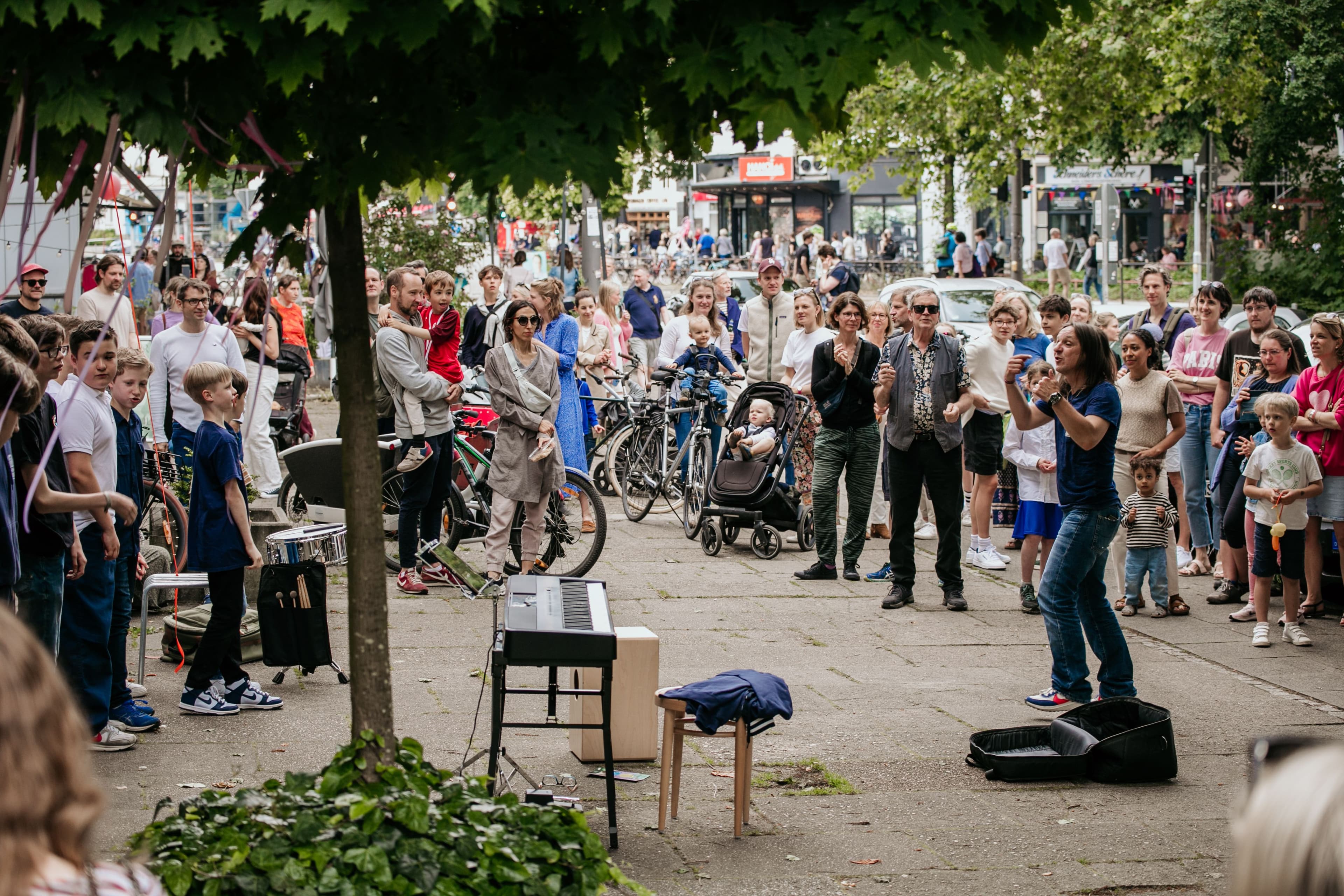 Viele Menschen stehen auf einer Straße um ein Keyboard herum. Eine Frau steht daneben und hat eine leere Gitarrentasche vor sich liegen.