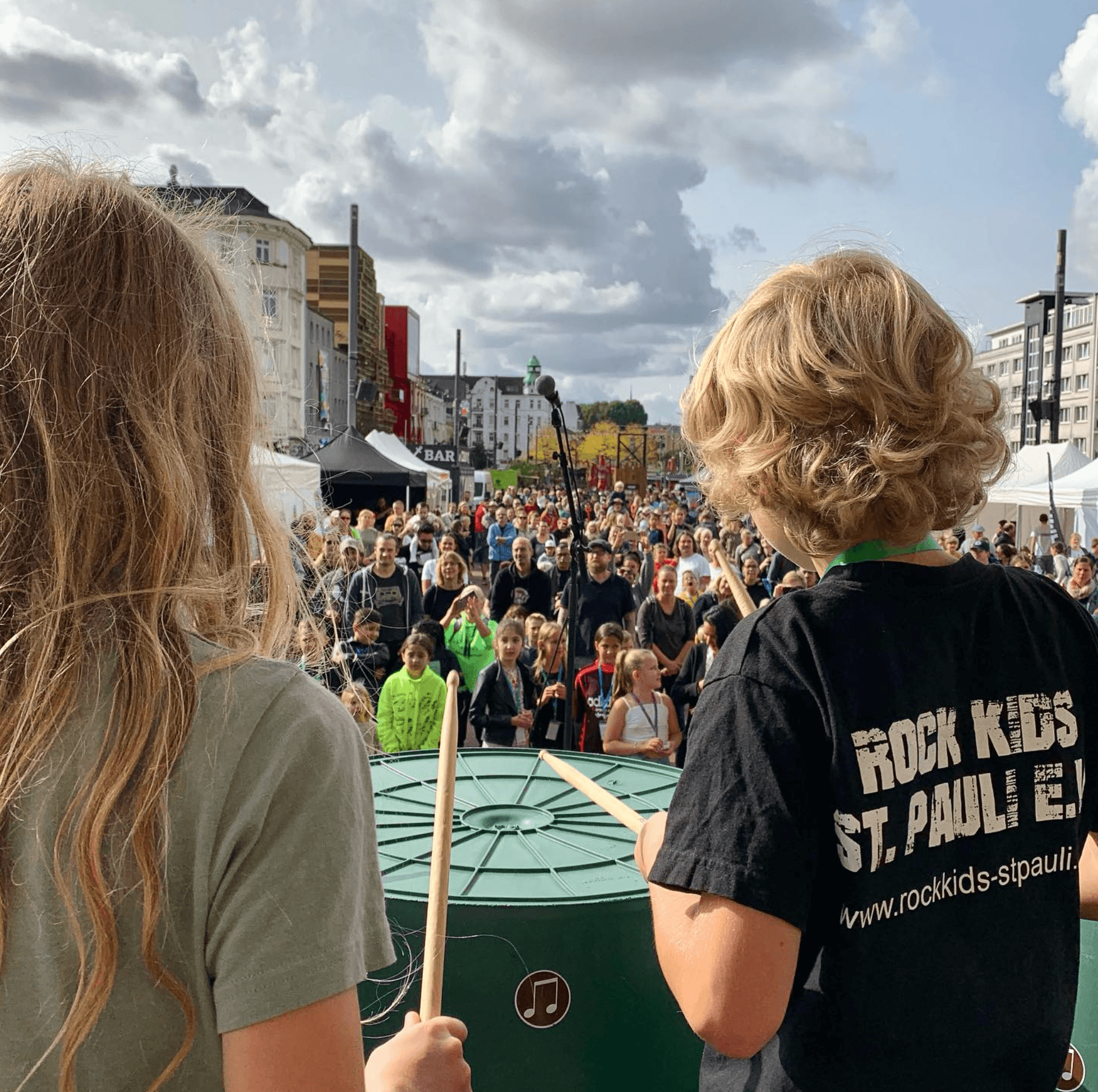 Zwei Kinder stehen mit Drumsticks in den Händen auf der Bühne, vor der Menschen sitzen und ihnen zuschauen