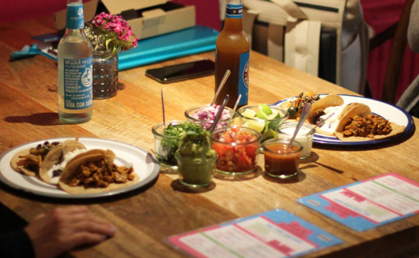 Auf einem Tisch stehen zwei Teller mit Tacos und in der Mitte des Tisches stehen einige Beilagen und Dips