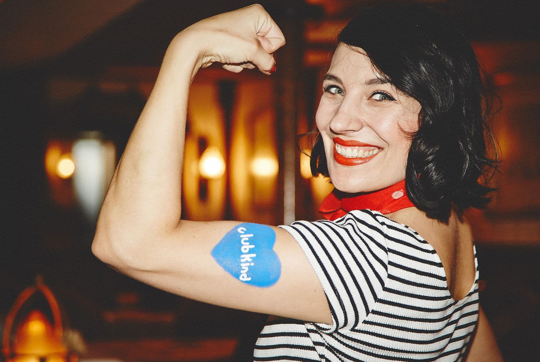 Eine Frau zeigt ihren Bizeps auf dem sie ein blaues clubkinder-Tattoo hat