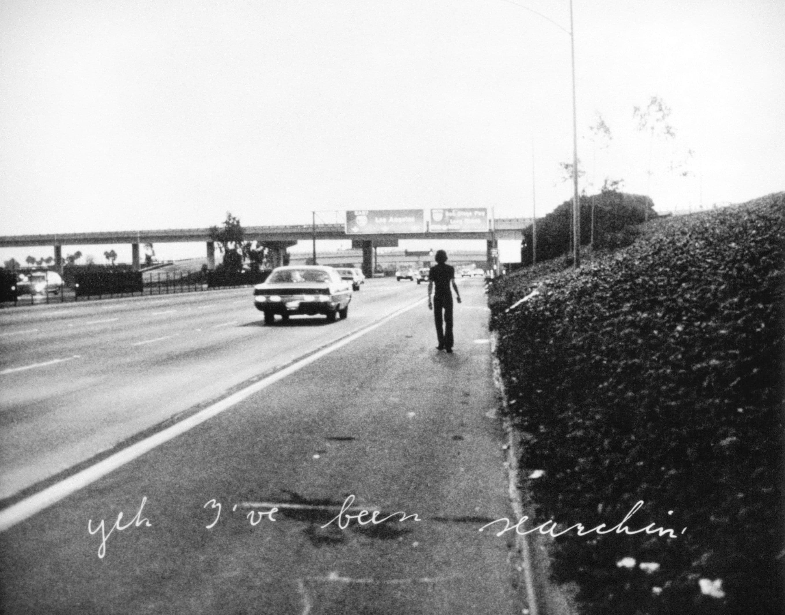 Bas Jan Aders Werk "In Search of the Miraculous" von 1973: Schwarz-weißes Foto einer Straße mit Autos und einem Fahrrad auf dem Gehweg. Auf dem Foto steht der Schiftzug "Yeh, I've been searching".