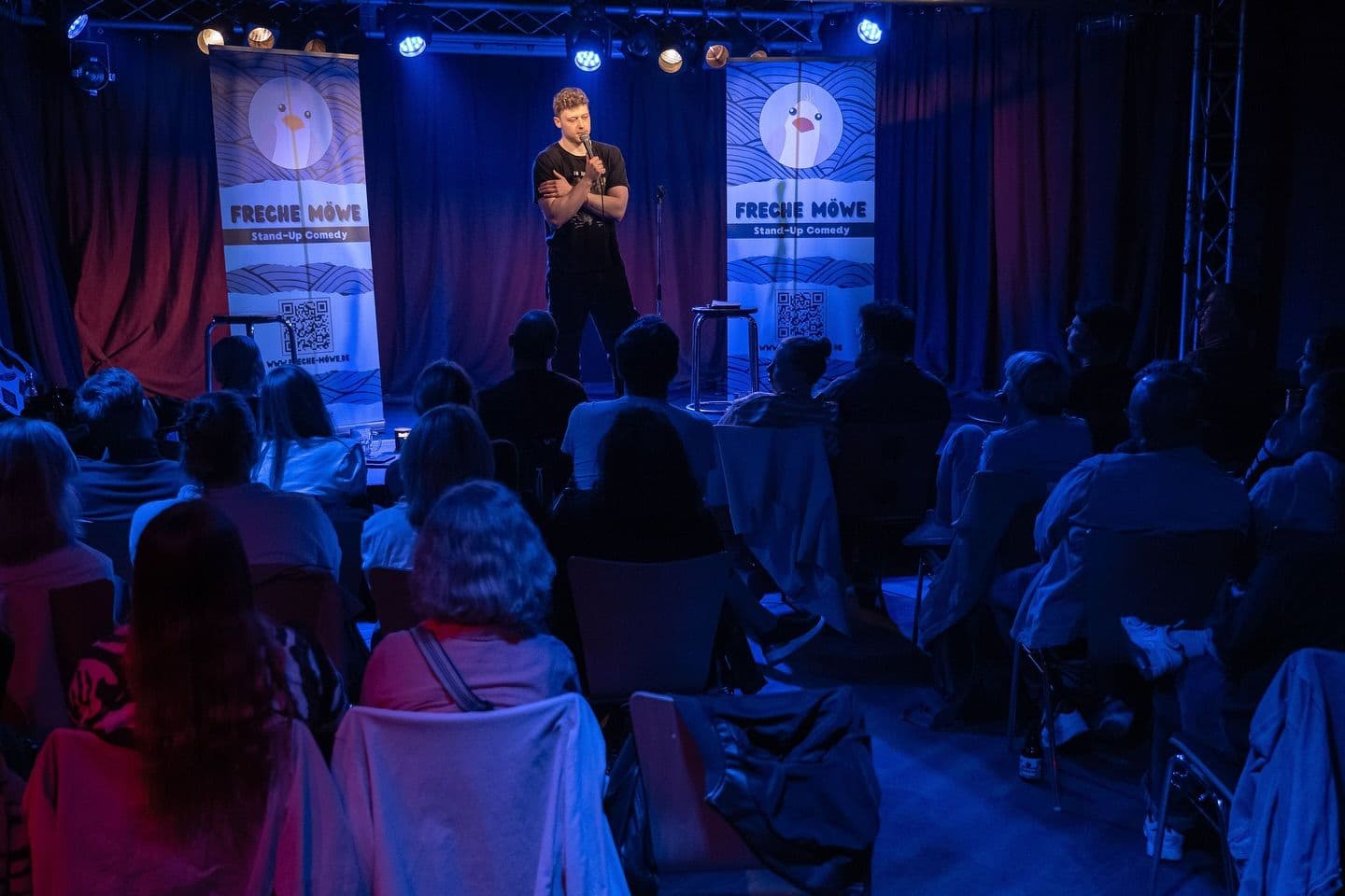 Ein Comedian steht mit einem Mikrofon auf der Bühne vor Publikum