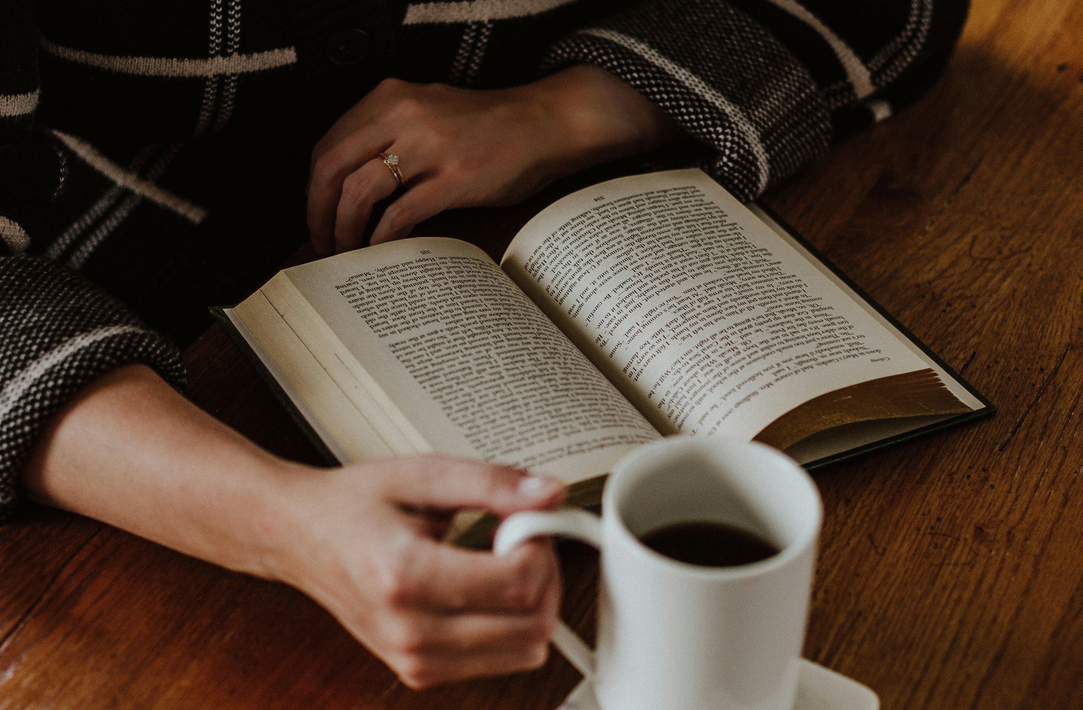 Eine Person sitzt vor einem aufgeschlagenen Buch mit einer Kaffeetasse in der Hand