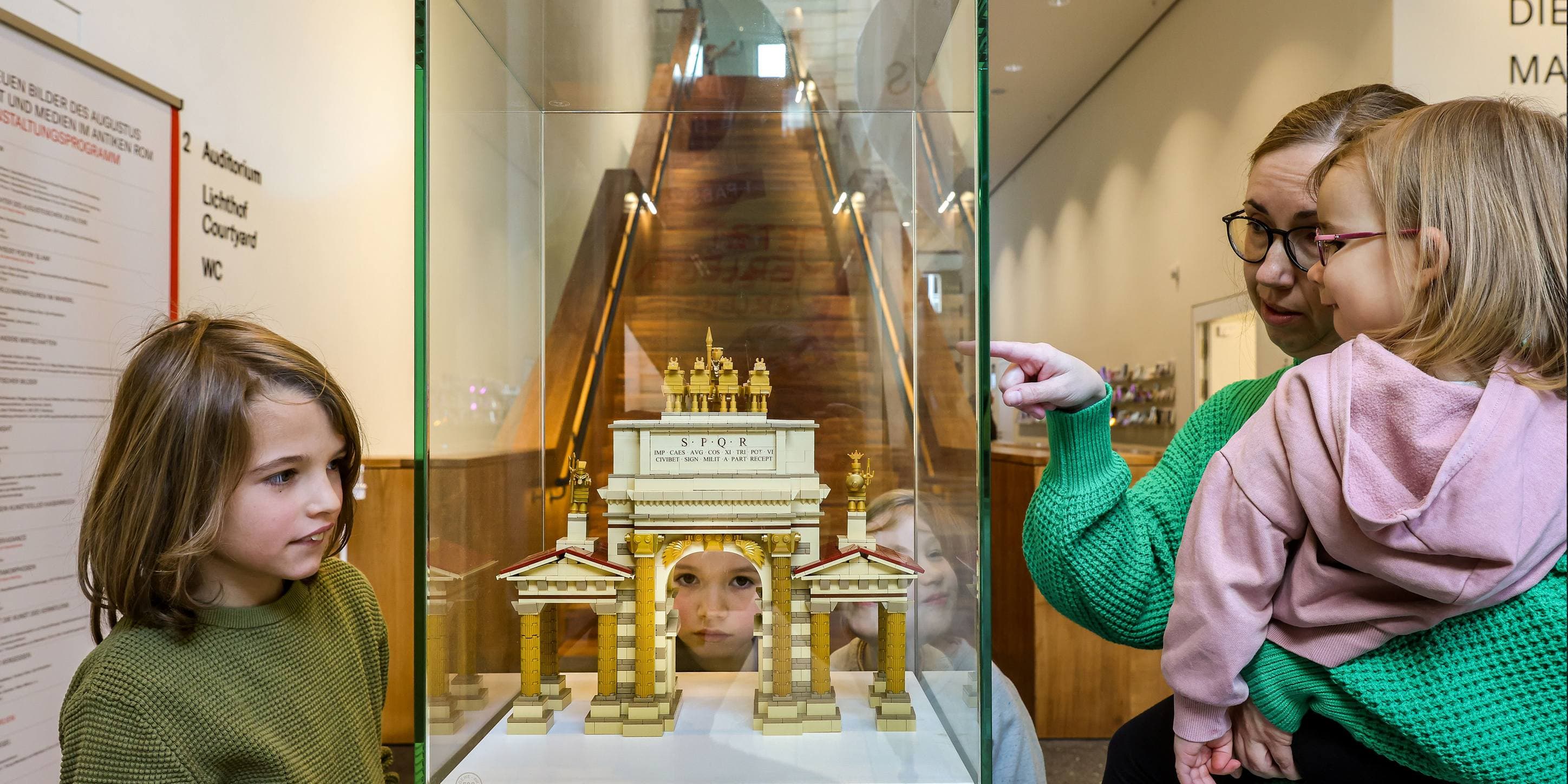 Eine Frau und zwei Kinder stehen an einem Glaskasten im Museum, in dem eine Lego-Nachbildung eines Bauwerks steht