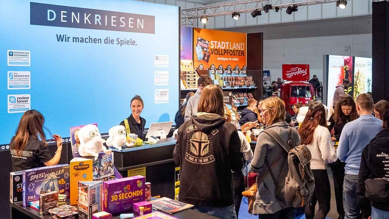 Eine Messehalle, in der ganz viele Menschen an Tischen sitzen und verschiedene Gesellschaftsspiele spielen. Im Hintergrund sind verschiedene Stände mit weiteren Spielen.