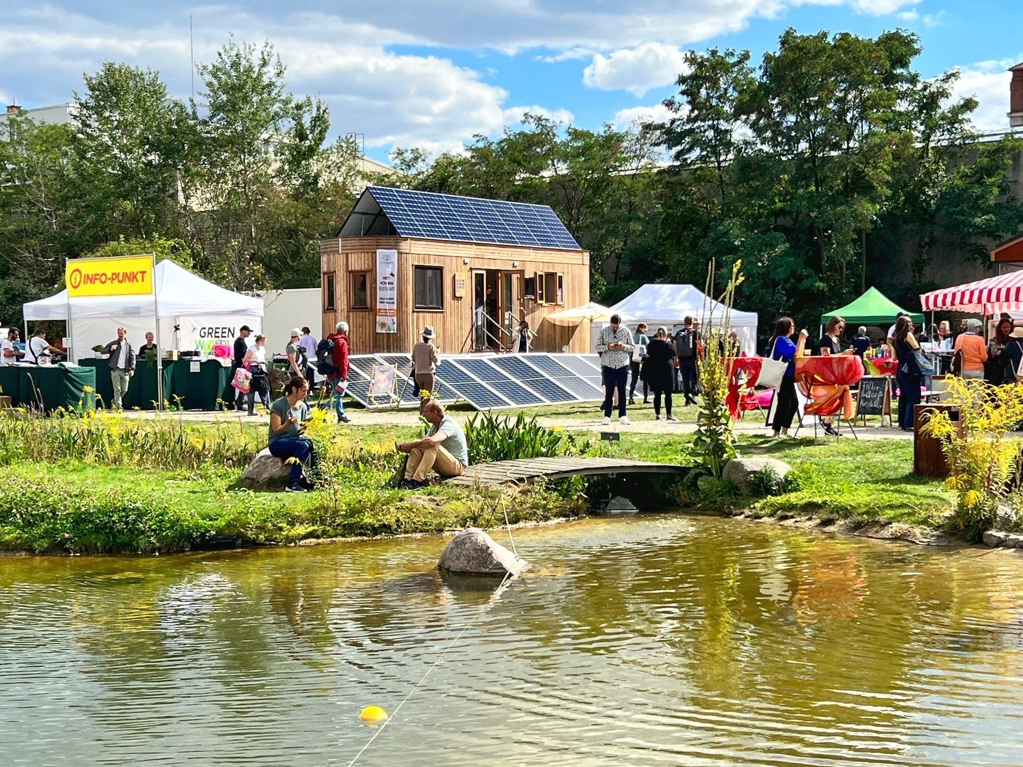 Übersicht über das Veranstaltungsgelände. Im Vordergrund ist ein See zu sehen, im Hintergrund Buden, Menschen und ein kleines Holzhaus.