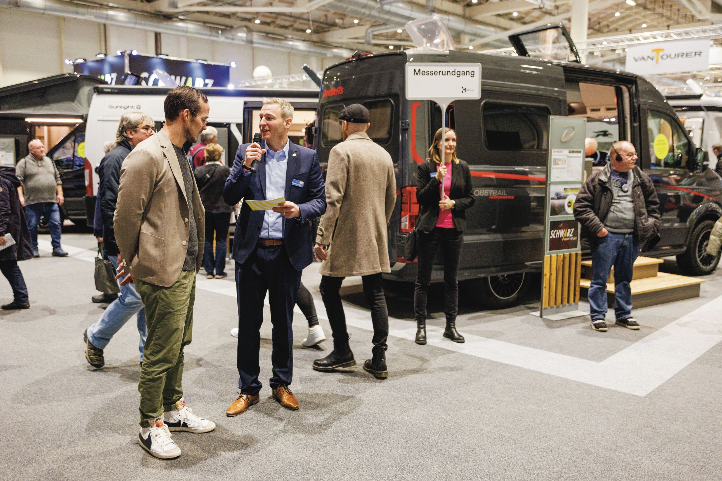 Ein Mann mit Mikrofon interviewt einen anderen Mann auf der Messe. Hinter ihm steht eine Frau mit einem "Messerundgang"-Schild in der Hand vor einem Caravan.