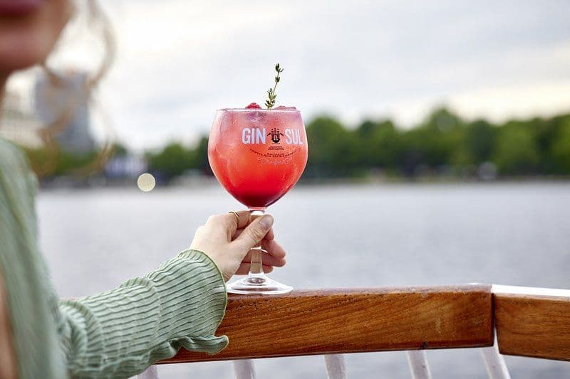 Person hält ein Gin Glas in die Kamera. Im Hintergrund liegt die Alster.