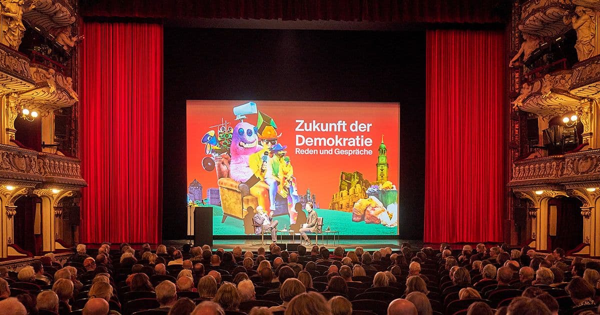 Ein Theaterpublikum blickt auf eine Bühne, auf der zwei Personen ein Gespräch führen.