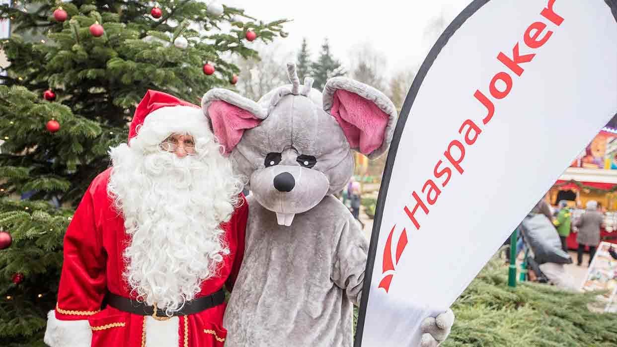 Die HaspaJoker Maus mit dem Weihnachtsmann vor einem geschmückten Tannenbaum