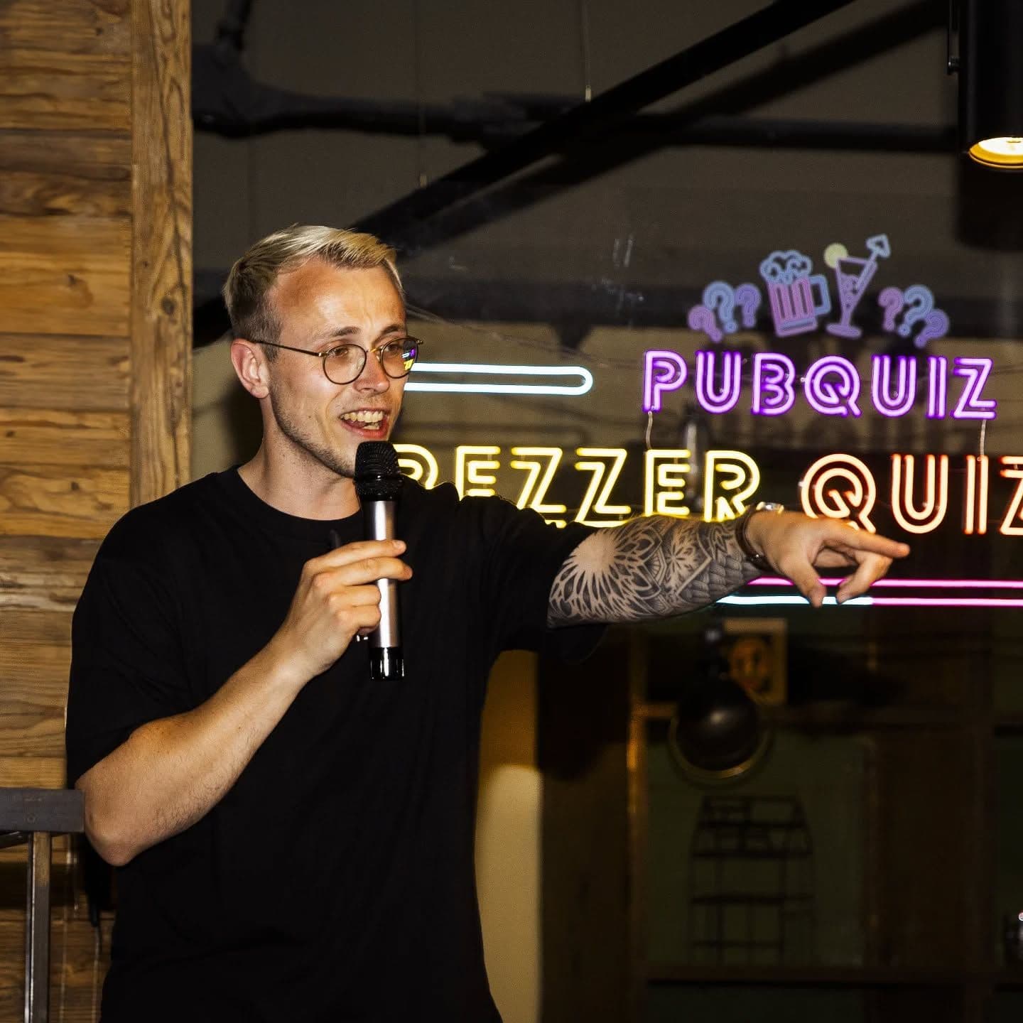 Ein Mann spricht in ein Mikrofon. Hinter ihm hängt ein LED-Schild mit der Aufschrift "Pubquiz Beezer Quizzer"