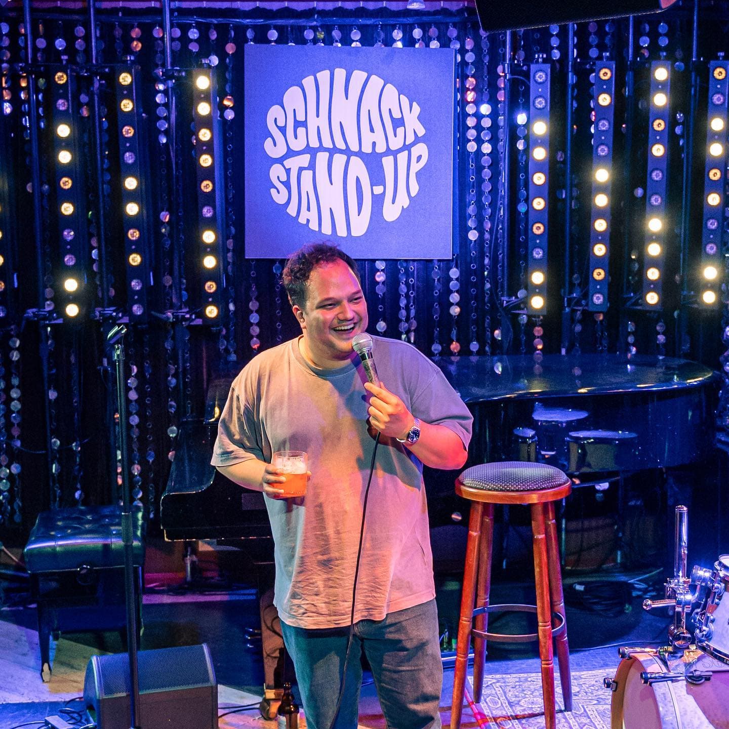Ein Mann steht mit einem Getränk in der einen und einem Mikrofon in der anderen Hand auf der Bühne. Hinter ihm hängt ein Schild mit der Aufschrift "Schnack Stand-Up".