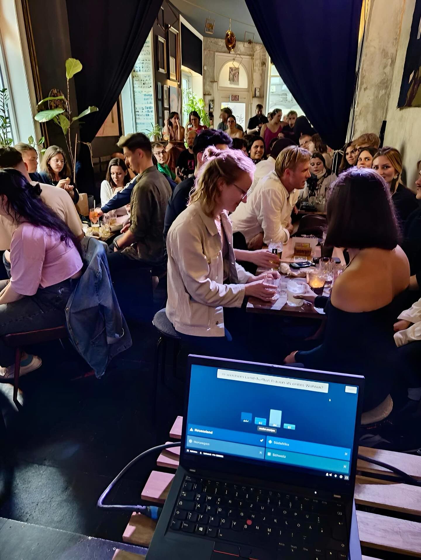 Menschen stehen an Bartischen und im Vordergrund steht ein Laptop auf einem Tisch